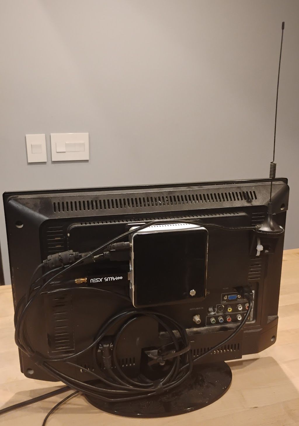 The back of a monitor. A small silver square is set against the monitor's vented black plastic case. An antenna is mounted on the right top corner and sticks up against a grey wall.